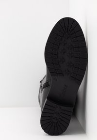 Semelle de botte noire avec des crampons profonds en motifs croisés, indiquant la taille 4,5 et la marque "Gabor" sur une surface blanche et un coin de mur.