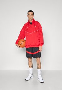 Rød Nike vindjakke med hvide detaljer og logo, sorte shorts med rød stribe, hvide sokker og basketballsko. Holder en basketball.
