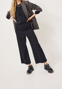 Pantaloni neri a gamba larga abbinati a una parte superiore scura e a un blazer grigio, completati da sneakers nere con accenti testurizzati.