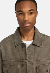 Olijfgroene denimjas met een knoopsluiting, twee borstzakken en een colkraag. Textuurstof met zichtbare stikseldetails.