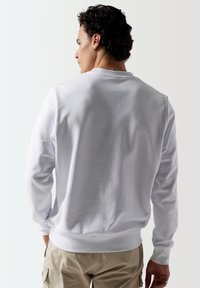 Sudadera de cuello redondo blanca con mangas largas, textura suave y dobladillos ribeteados, mostrada desde la parte de atrás.