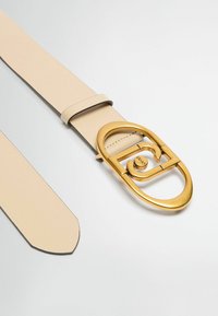 Cintura in pelle beige con una grande fibbia ovale in oro lucido che presenta un design intrecciato e un elemento centrale arrotondato. I bordi sono lisci.