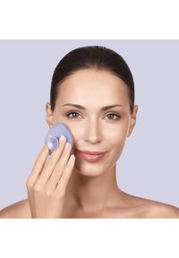Dispositivo per la pulizia del viso in silicone, lilla, di forma rotonda con superficie testurizzata, utilizzato sulla pelle del viso di una persona. Carnagione liscia.