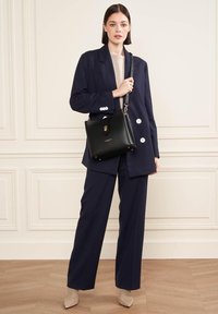 Borsa a tracolla in pelle nera con una chiusura dorata, abbinata a un blazer scuro, una maglietta chiara e pantaloni a gamba larga. Tacchi neutri completano l'outfit.