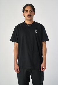 Schwarzes Baumwoll-T-Shirt mit rundem Halsausschnitt, kurzen Ärmeln und einem kleinen weißen gestickten Design auf der linken Brust.