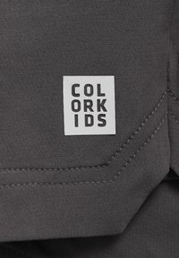 Szürke szövet sima textúrával, varrott széllel, amelyen egy fehér téglalap alakú címke található, rajta a "COLOR KIDS" felirat vastag fekete betűkkel.