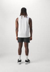 Chemise sans manches blanche avec une texture lisse, associée à un short noir. Le modèle porte des chaussettes blanches et des chaussures de sport noires.