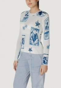 Maglione bianco con stampe grafiche blu, tra cui piante ed etichette decorative. Le maniche lunghe e l'orlo a coste aggiungono texture.