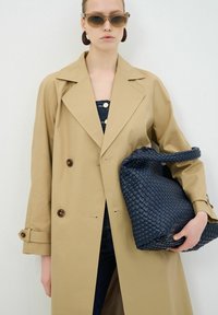 Beige trenchcoat met dubbele knopen en brede kraag, gedragen over donkere denim. Het model houdt een grote marineblauwe geweven handtas vast met een stevige hengsel.