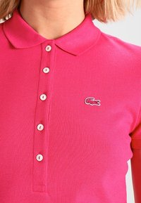 Polo shirt rosa realizzata in tessuto testurizzato. Presenta un colletto classico, una chiusura con cinque bottoni e un piccolo logo ricamato sul petto.