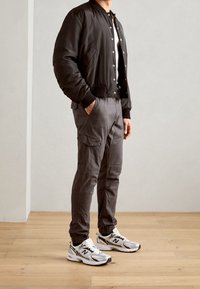 Veste bomber noire avec boutons-pression, pantalon cargo gris avec poches, et baskets blanches à textures multiples avec accents noirs.