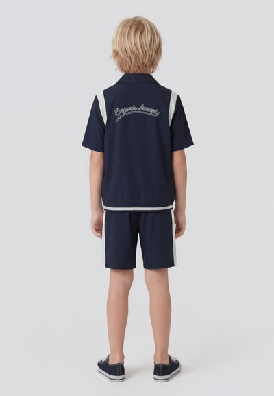 Παιδί με ξανθά μαλλιά που κοιτάζει προς τα πίσω, φορώντας μπλε σκούρο κοντομάνικο μπουφάν και σορτς με λευκές ρίγες, μπλε σκούρα αθλητικά παπούτσια, "Emporio Armani" στο πίσω μέρος του μπουφάν.