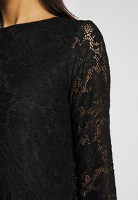 Gros plan d'un haut en dentelle noire pour femme avec des motifs floraux et des manches longues semi-transparentes.