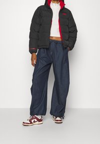 Zwarte pufferjas met rode voering, witte cropped top, losse blauwe denim broek en bordeauxrode en witte sneakers. De jas heeft een logodetail.