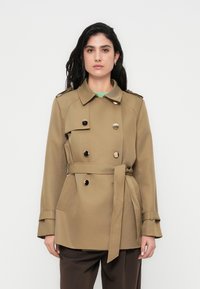 Μπεζ διπλό παλτό τύπου trench με ζώνη, κουμπιά διακοσμητικά, επιβραχίονες και πλαϊνές τσέπες, συνδυασμένο με σκούρα καφέ παντελόνια.