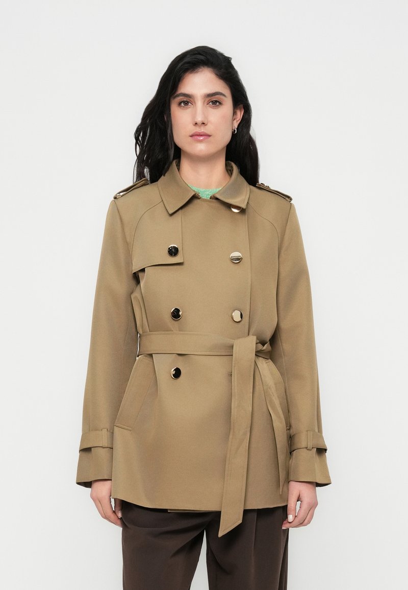 Μπεζ διπλό παλτό τύπου trench με ζώνη, κουμπιά διακοσμητικά, επιβραχίονες και πλαϊνές τσέπες, συνδυασμένο με σκούρα καφέ παντελόνια.