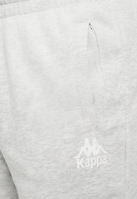 Hellgraue Jogginghose mit Seitentaschen, aus weichem Stoff hergestellt, mit einem weißen Kappa-Logo, das auf der unteren linken Seite gedruckt ist.