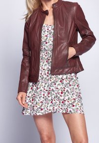 Mujer vestida con una chaqueta de cuero burdeos sobre un vestido floral blanco con flores rosas, rojas y amarillas, de pie con una mano en el bolsillo de la chaqueta.