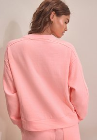 Pink textured sweatshirt with a boxy shape, ribbed accents at shoulders, and a slightly cropped hem. Smooth fabric with a relaxed fit.