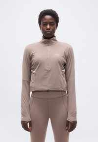 Άτομο που κοιτάει προς τα εμπρός, φοράει τοπ με φερμουάρ σε χρώμα ταουπέ με μακριά μανίκια και ψηλόμεσα κολάν στο ίδιο χρώμα, μπροστά σε απλό ανοιχτόχρωμο φόντο.