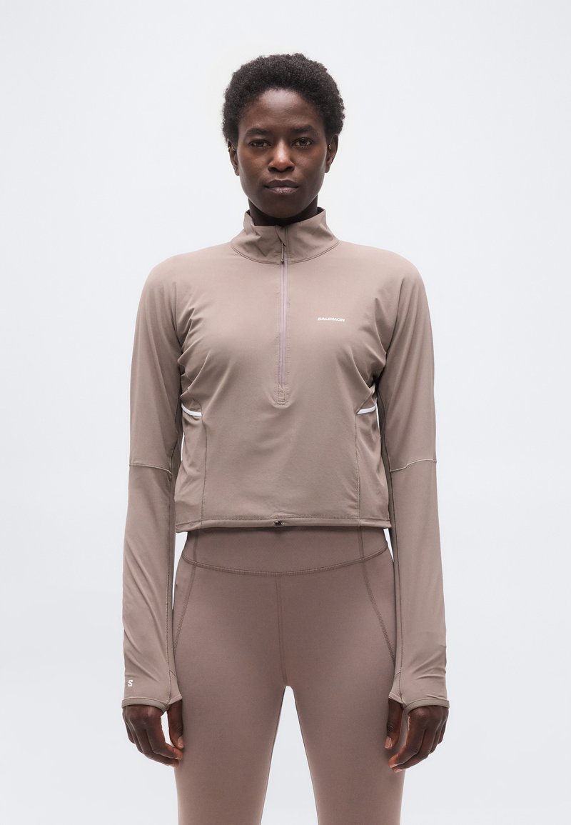 Άτομο που κοιτάει προς τα εμπρός, φοράει τοπ με φερμουάρ σε χρώμα ταουπέ με μακριά μανίκια και ψηλόμεσα κολάν στο ίδιο χρώμα, μπροστά σε απλό ανοιχτόχρωμο φόντο.