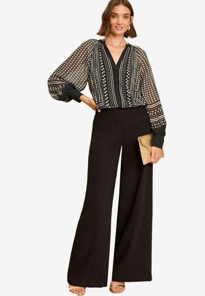 Kobieta ubrana w czarną i białą bluzkę w groszki z przezroczystymi, balonowymi rękawami, czarne szerokie spodnie oraz nude sandały, trzymająca złotą kopertówkę.