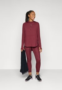 Vinröd träningsöverdel med huva och ficka, kombinerad med mönstrade vinröda leggings. Modellen håller en svart handduk och bär svarta träningsskor.