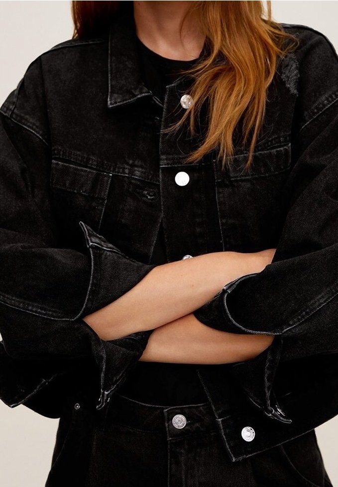 Personne aux longs cheveux roux portant une veste et un pantalon en jean noir, bras croisés, sur un fond clair uni.