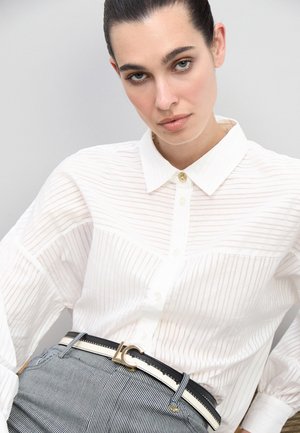 Camisa blanca de botones con rayas verticales, con cuello clásico y botón dorado. Combinada con un cinturón a rayas en blanco y negro.