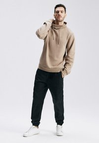 Beżowa bluza z kapturem, z sznurkiem, z ribbowymi mankietami i dołem; zestawiona z czarnymi spodniami dresowymi i białymi sneakersami. Minimalistyczny design.