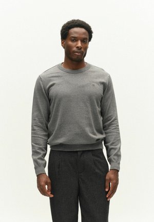 Man wearing a light gray long-sleeve sweater and dark gray pants standing against a plain white background.