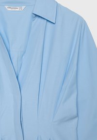 Chemise bleu clair à manches longues avec col, devant ouvert et étiquette visible à l'intérieur indiquant Stradivarius, taille EUR S, fabriquée en Inde.