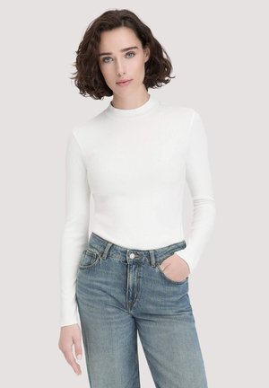 Witte ribgebreide longsleeve top met een hoge hals, gedragen met lichtblauwe relaxed-fit jeans. Het model staat tegen een neutrale achtergrond.