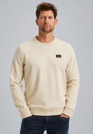 Beige sweatshirt with a round neck and ribbed cuffs. Features a small black logo patch on the chest and a smooth cotton texture.