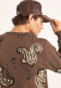 Suéter de punto marrón con un patrón de paisley en crema y verde, combinado con una gorra de béisbol marrón de malla. Tela texturizada visible.
