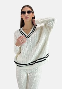 Suéter de punto blanco con patrón de trenzas, cuello en V y detalles de rayas negras en el escote y el dobladillo, combinado con pantalones de punto trenzado a juego.