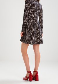 Kvinna som bär en svart klänning med vita prickar och röda högklackade ankelskor, stående mot en enkel bakgrund.