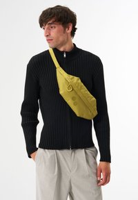 Gelbe, wasserabweisende Umhängetasche mit flachem Design und Reißverschluss, die zwei runde Akzente und einen verstellbaren Riemen bietet.