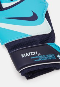Marinblå och akvamarinfärgade målvaktshandskar med texturerat grepp, med en Nike-logotyp och etikett "MATCH JR". Materialet är andningsbart tyg med vaddering.