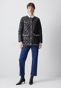 Quilted black jacket with two front pockets, white collar, gold buttons, and textured trim. Paired with blue denim pants and black heeled boots.