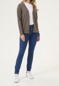 Brauner leichter Cardigan über einem weißen T-Shirt, kombiniert mit dunkelblauen Skinny-Jeans und weißen Sneakern, steht vor einem hellen Hintergrund.