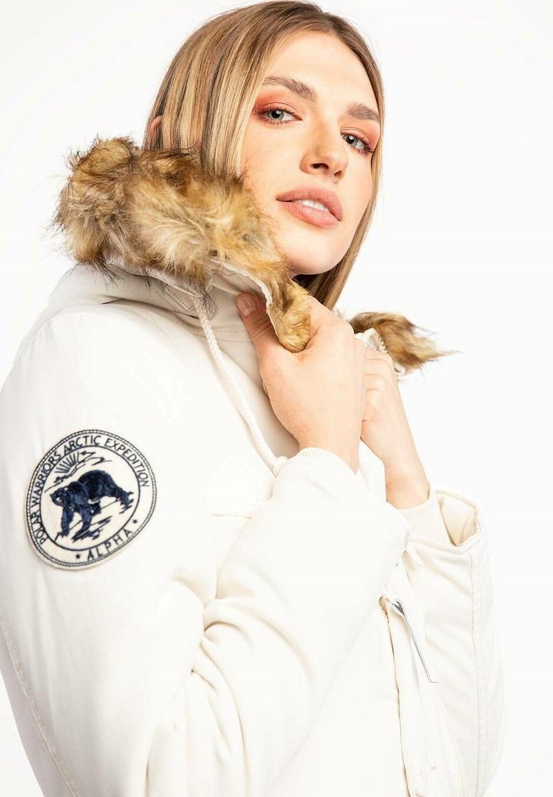 Frau, die die Kapuze einer weißen Winterjacke mit Fellbesatz und einem Arctic Expedition-Patch am Ärmel anpasst, schaut mit neutralem Ausdruck in die Kamera.