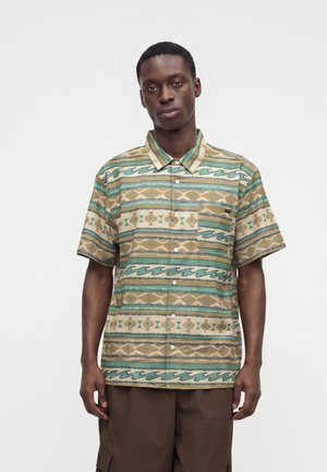 Homme portant une chemise à manches courtes boutonnée avec des motifs tribaux horizontaux beige et verts et un short cargo marron, debout devant un fond uni.