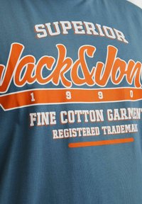 Camiseta de algodón azul con texto en negrita blanco y naranja, que incluye "SUPERIOR," "Jack & Jones," y "FINE COTTON GARMENT," con detalles de marca registrada.