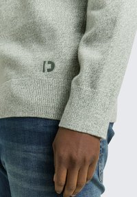 Lichtgrijze, gestructureerde trui met een geribbelde zoom en manchetten, voorzien van een klein groen logo bij de tailleband, gecombineerd met donkerblauwe jeans.