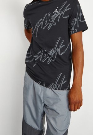 Hombre vistiendo una camiseta negra de Jordan Flight con texto gráfico y pantalones cortos gris claro frente a un fondo liso.