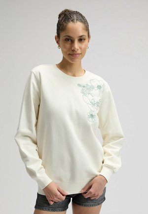 Giovane donna con i capelli raccolti in uno chignon, indossa una felpa color crema con ricamo floreale blu sulla spalla e shorts di denim scuro in piedi.