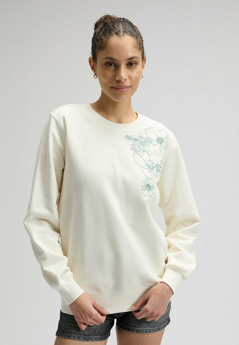 Joven con el cabello recogido en un moño, vistiendo una sudadera crema con bordado floral azul en el hombro y shorts de mezclilla oscuros, de pie.