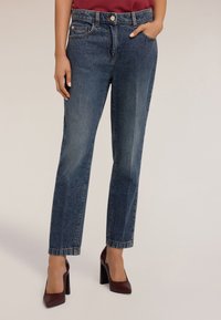 Pantalones vaqueros de mezclilla azul oscuro con corte recto, cinco bolsillos y cierre de botón. Combinados con zapatos de tacón cuadrado color burdeos. Textura suave.