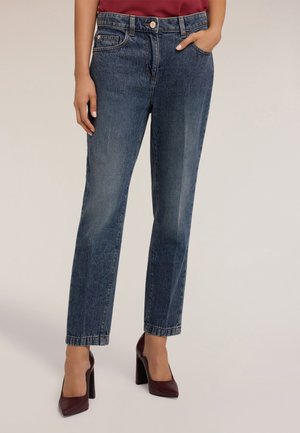 Pantalones vaqueros de mezclilla azul oscuro con corte recto, cinco bolsillos y cierre de botón. Combinados con zapatos de tacón cuadrado color burdeos. Textura suave.